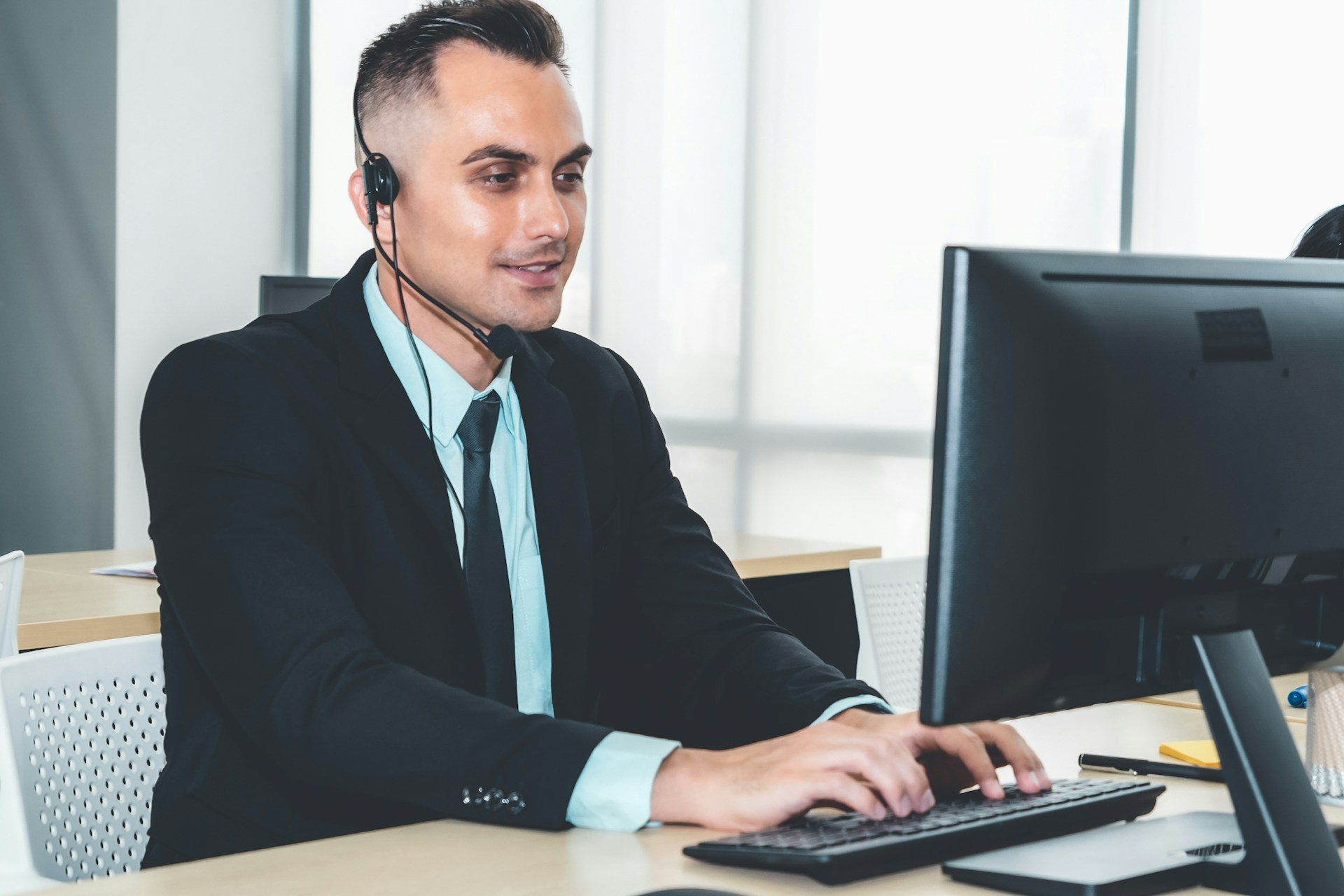 Man in a suit and headset works on a computer at a modern office desk.